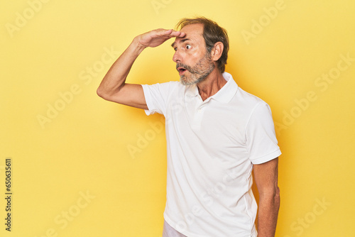 Caucasian middle-aged man in yellow studio looking far away keeping hand on forehead.