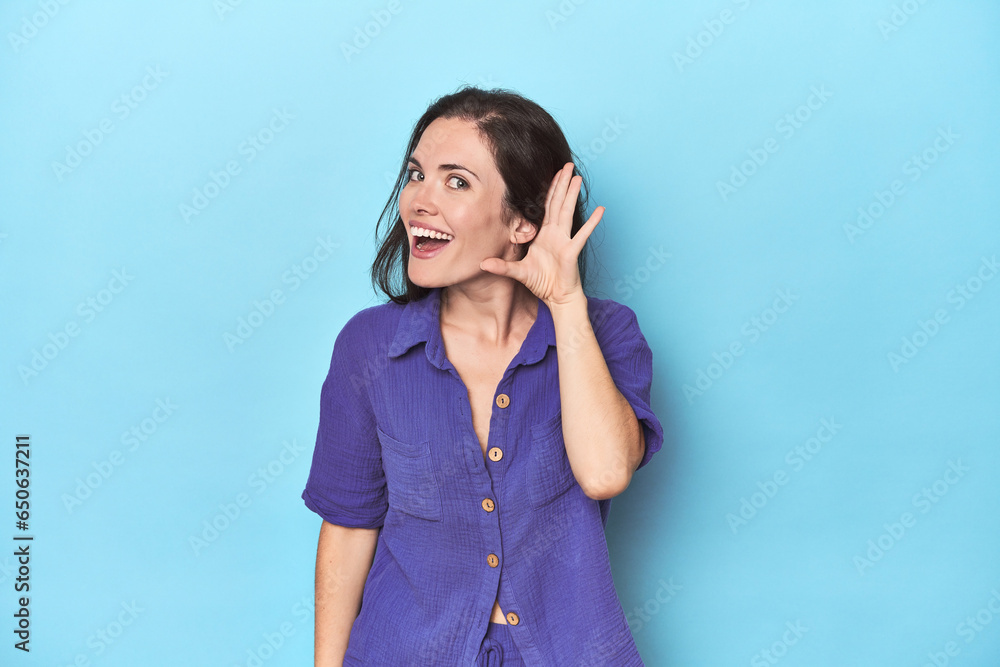 Young caucasian woman on blue backdrop trying to listening a gossip.
