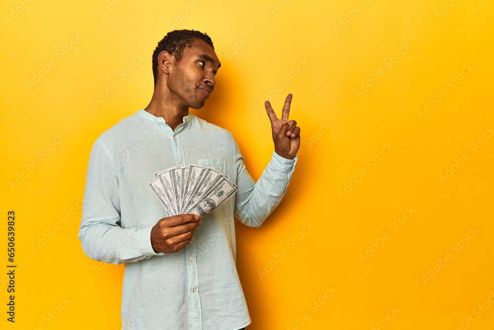 African American man with dollar bills, yellow studio, joyful and carefree showing a peace symbol with fingers. African American man with dollar bills, yellow studio, joyful and carefree showing a peace symbol with fingers.