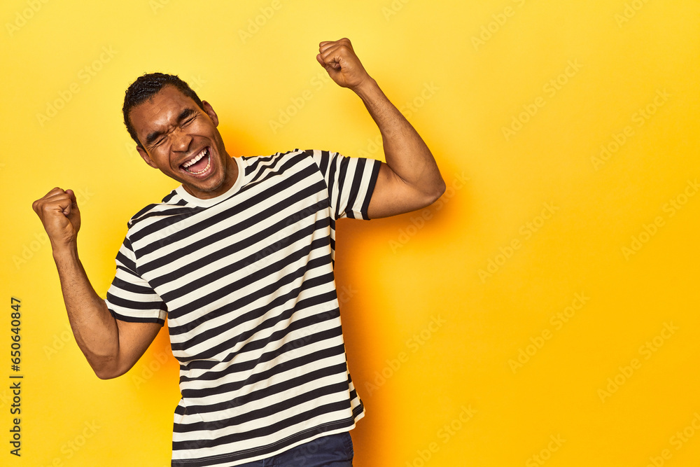 African American man in striped t-shirt, yellow studio, raising fist after a victory, winner concept. African American man in striped t-shirt, yellow studio, raising fist after a victory, winner concept.