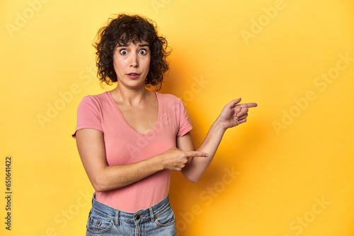 Curly-haired Caucasian woman in pink t-shirt shocked pointing with index fingers to a copy space.