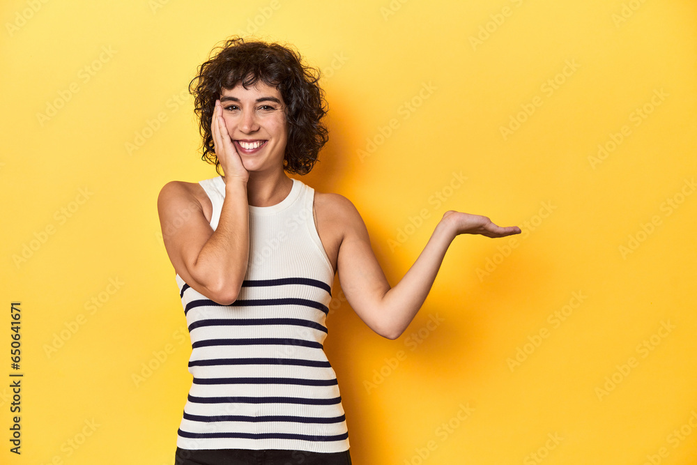 Caucasian curly-haired woman in white tank-top holds copy space on a palm, keep hand over cheek. Amazed and delighted. Caucasian curly-haired woman in white tank-top holds copy space on a palm, keep hand over cheek. Amazed and delighted.