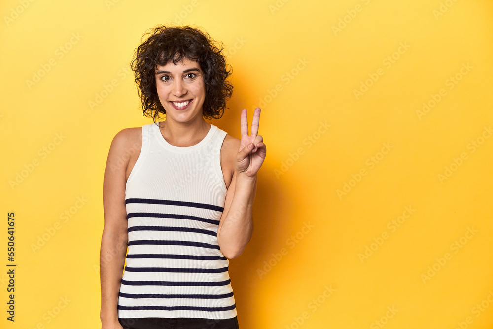 Caucasian curly-haired woman in white tank-top showing victory sign and smiling broadly. Caucasian curly-haired woman in white tank-top showing victory sign and smiling broadly.