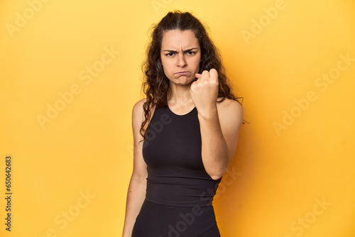 Принти на полотні Sporty woman in active wear, yellow backdrop, showing fist to camera, aggressive facial expression
