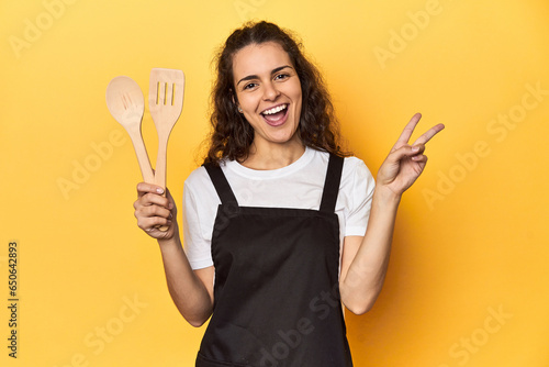 Woman with apron, wooden cooking utensils, yellow, joyful and carefree showing a peace symbol with fingers.