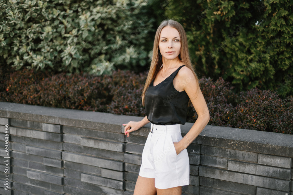 A girl with long blond hair, wearing a black top, white shorts, poses against a background of a gray wall. Young woman walking through a park with modern architecture and landscape design A girl with long blond hair, wearing a black top, white shorts, poses against a background of a gray wall. Young woman walking through a park with modern architecture and landscape design