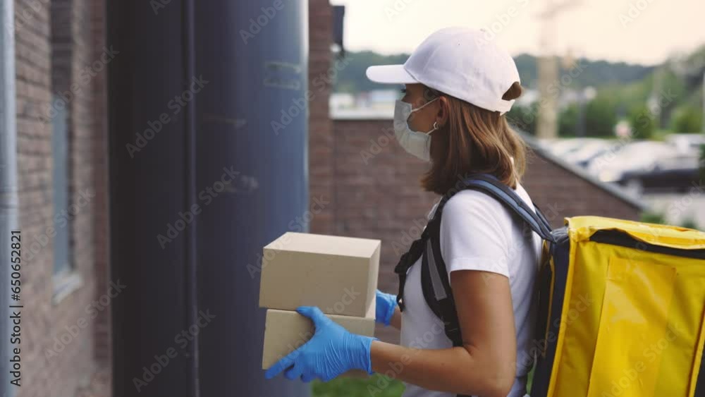 A delivery female courier wearing a protective mask and gloves, delivers a paper boxes, wearing a medical mask, safety gloves, and a uniform. Delivering woman calling the intercom A delivery female courier wearing a protective mask and gloves, delivers a paper boxes, wearing a medical mask, safety gloves, and a uniform. Delivering woman calling the intercom