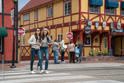 full length of two asian japanese female visitors discussing landmark at distance with pointing gesture as they are crossing main street of solvang lined up with colorful Danish architectures