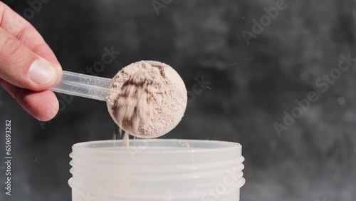 Male hand puts into a shaker portion of chocolate whey protein powder from measuring scoop on dark background, slow motion, close-up view