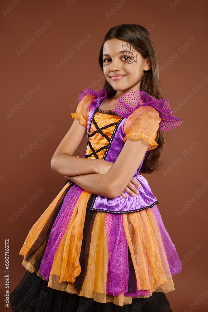 happy kid in Halloween costume with spiderweb makeup standing with folded arms on brown backdrop