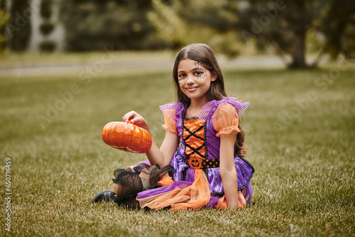 joyful girl in Halloween costume sitting on green grass and holding decorative pumpkin photo