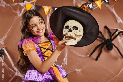 happy girl in Halloween attire wearing witch hat on skull on brown background, spooky season photo