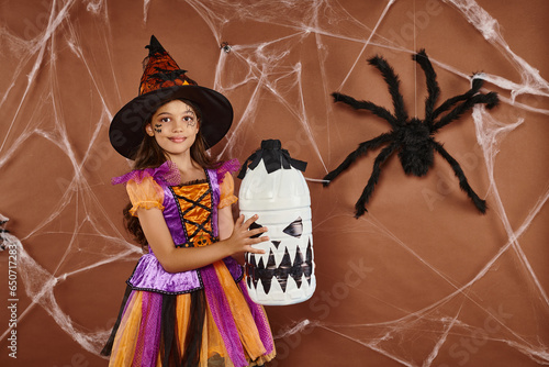 happy girl in witch hat and Halloween costume standing with spooky decor on brown background photo