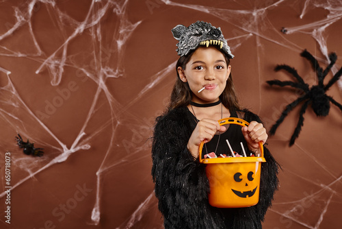 close up girl with lollipop holding candy bucket on brown backdrop with spiderweb, Halloween photo