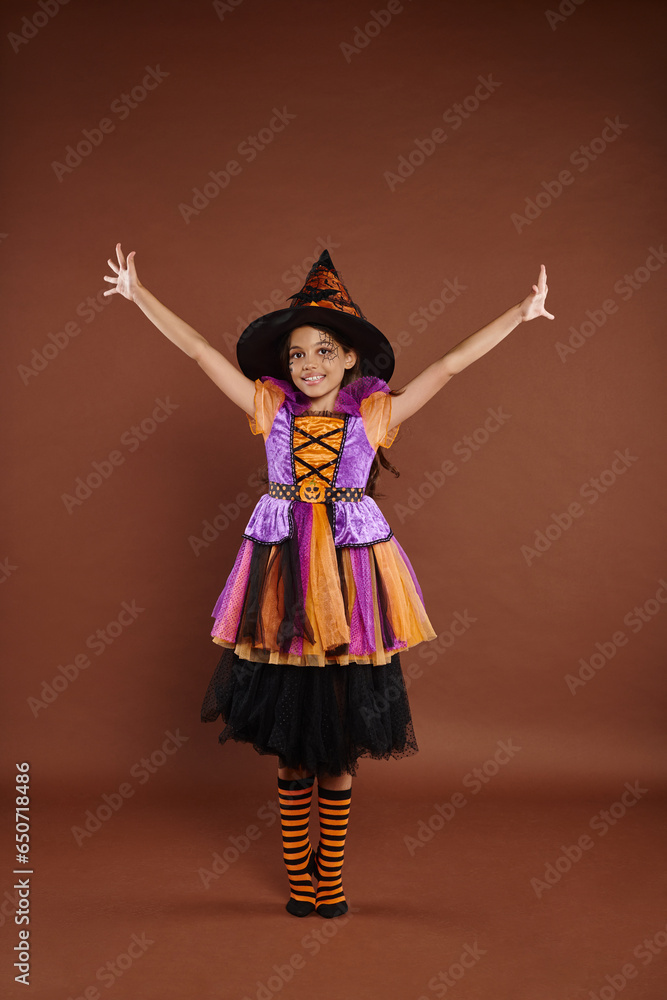 excited girl in Halloween costume and pointed hat standing with raised hands on brown background excited girl in Halloween costume and pointed hat standing with raised hands on brown background