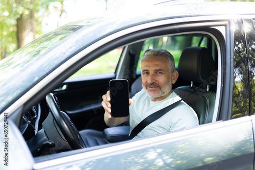 Mobile app for car rental, car parking. Cheerful man sitting in car, showing cell phone with white blank screen