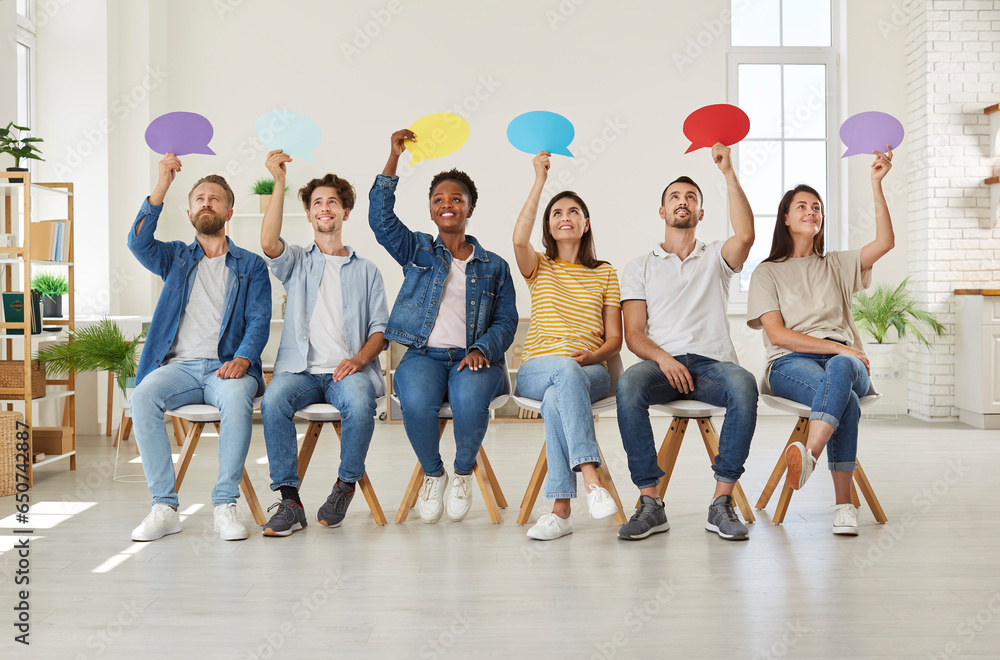 Obraz premium Group of happy young diverse multiracial people holding up colorful paper mockup speech bubbles while sitting in row on chairs in new modern office or rented apartment. Opinion survey concept