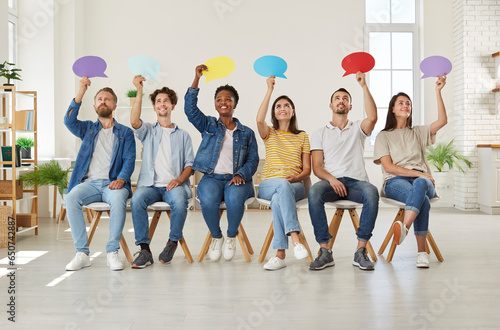 Group of happy young diverse multiracial people holding up colorful paper mockup speech bubbles while sitting in row on chairs in new modern office or rented apartment. Opinion survey concept