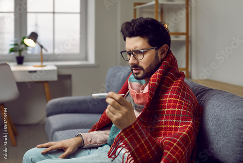 Fototapeta Sick bearded young man in glasses and a blanket sitting on the couch at home and looking at the thermometer in his hand