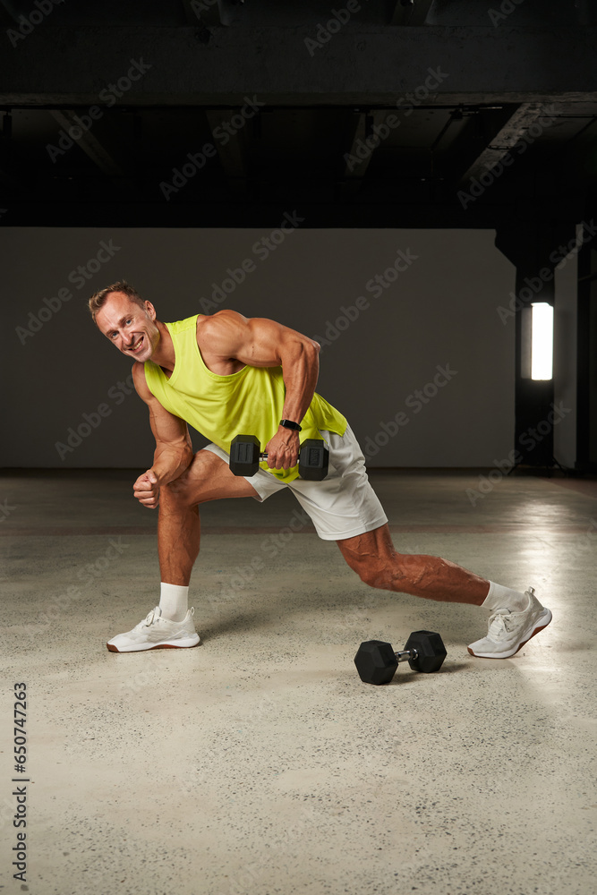 a handsome smiling Caucasian athlete is engaged in a sprint with dumbbells. lunges onto the knee with a dumbbell in his hand. he is wearing white shorts and a yellow T-shirt