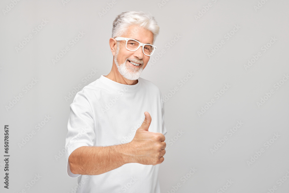 Portrait of mature man in white T-shirt and glasses smiling joyfully showing thumbs up gesture isolated on white studio background. Approves good choice, right decision. Portrait of mature man in white T-shirt and glasses smiling joyfully showing thumbs up gesture isolated on white studio background. Approves good choice, right decision.