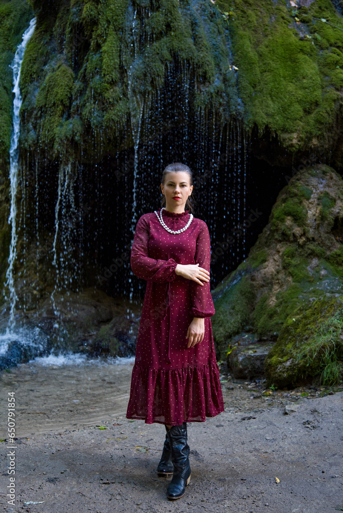 beautiful romantic brunette woman in a vintage burgundy dress with red lips and white necklace standing near a waterfall at sunset in autumn