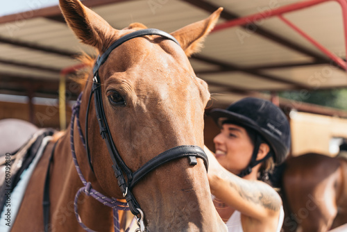 Young jockey smiling next to his horse ready to ride. Middle-aged woman finishing the preparations of her equestrian animal for her riding lesson.
