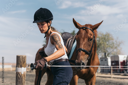 Young jockey inside the training enclosure of the equestrian center with his horse. Middle-aged woman closing the security cord that closes the equestrian training arena. © Jorge