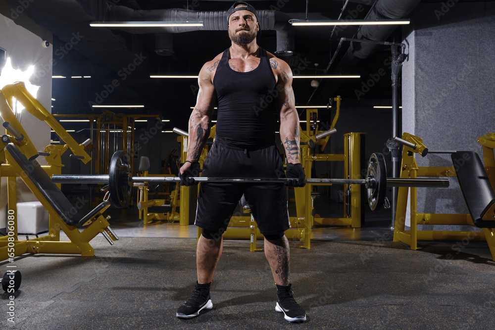 Bearded athlete in black sport gloves, shorts, vest, cap and sneakers. He lifting a barbell, posing in dark gym with yellow equipment. Full length Bearded athlete in black sport gloves, shorts, vest, cap and sneakers. He lifting a barbell, posing in dark gym with yellow equipment. Full length