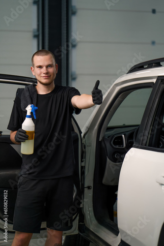 Portrait of a young car wash worker holding a spray gun and showing a thumbs up sign on the background of a white SUV in the process of detangling