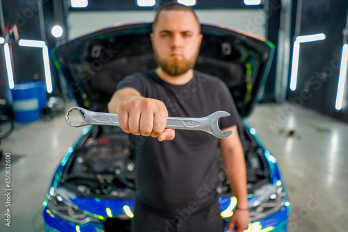 Portrait of a serious car service worker standing with a wrench in his hand against the background of a chameleon-colored car with an open hood