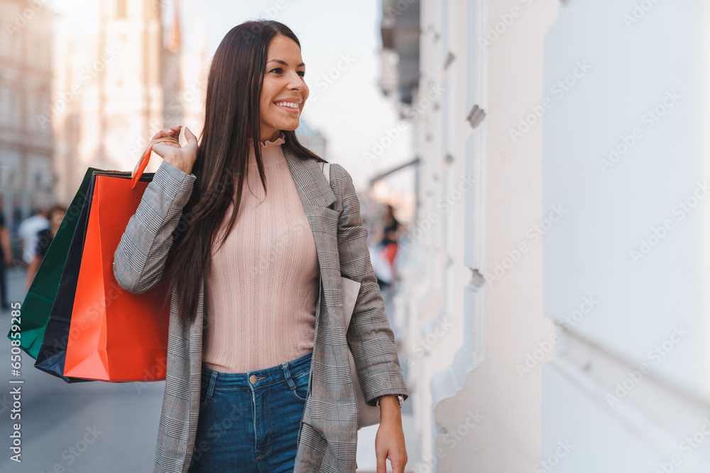 Attractive young woman with long well-groomed brown hair smiling and looking at store window while holding shopping bags over shoulder on the city street.