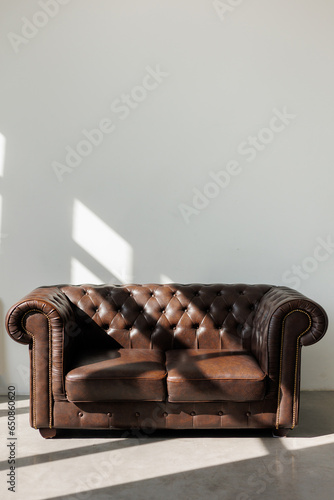 Vintage quilted classic brown sofa against white wall with sunlights from window on concrete floor at studio. Natural leather with rhombic stitching with buttons. Furniture design. Copy space
