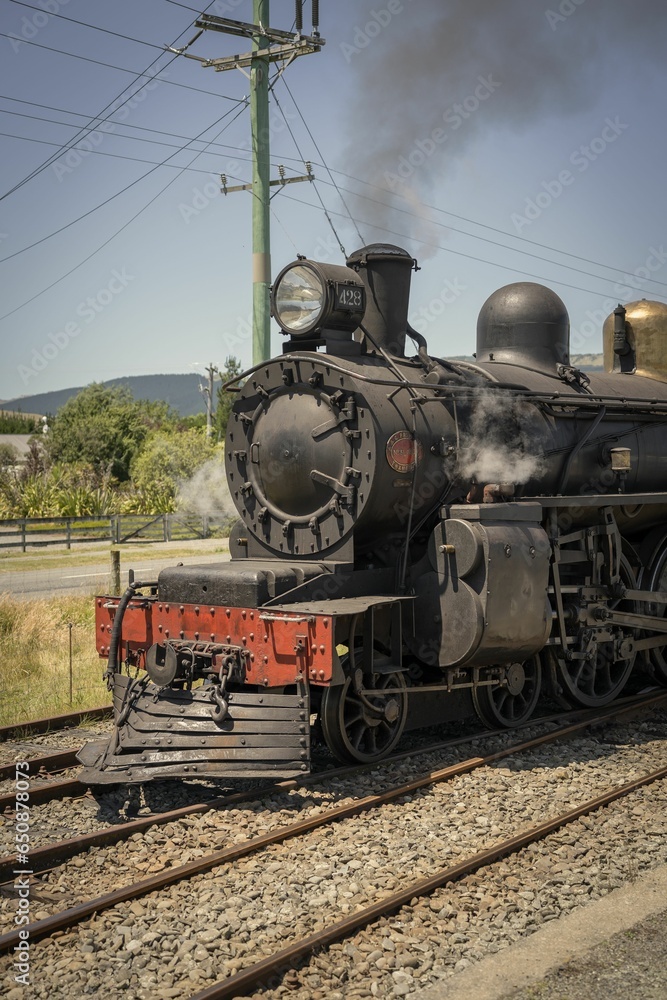 Obraz premium Aged, corroded train engine travels along a track in a train yard with a plume of smoke