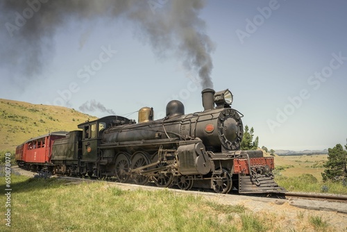 Aged, corroded train engine travels along a track in a train yard with a plume of smoke