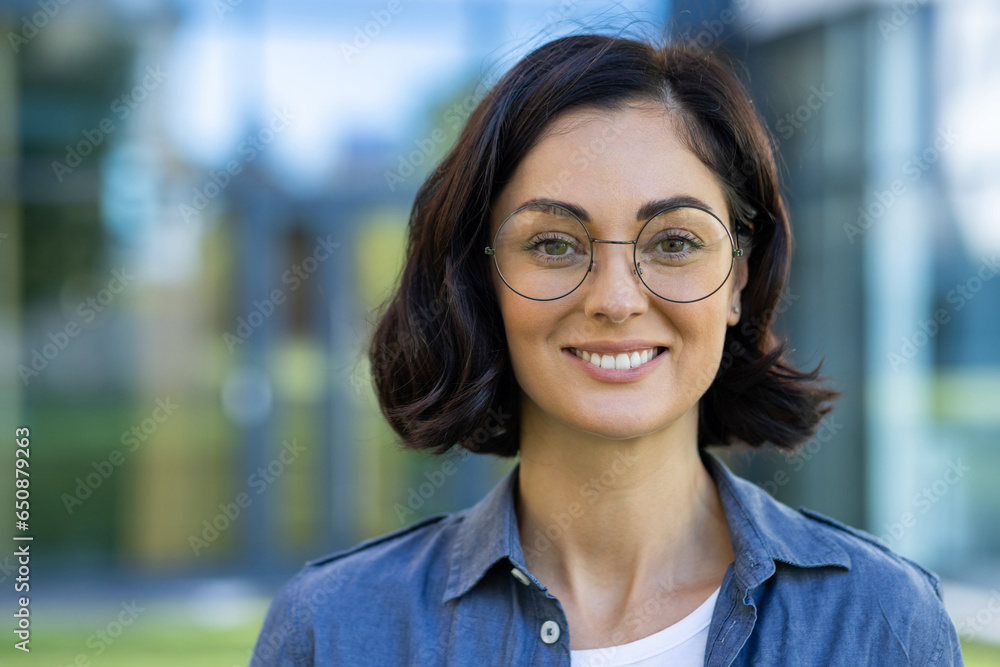 Close-up portrait of a young beautiful woman in glasses and a shirt, a student smiling and looking at the camera outside a university campus on a sunny day Close-up portrait of a young beautiful woman in glasses and a shirt, a student smiling and looking at the camera outside a university campus on a sunny day