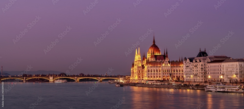 Fototapeta premium Panoramic view of illuminated grand buildings at the shore near a bridge at sunset