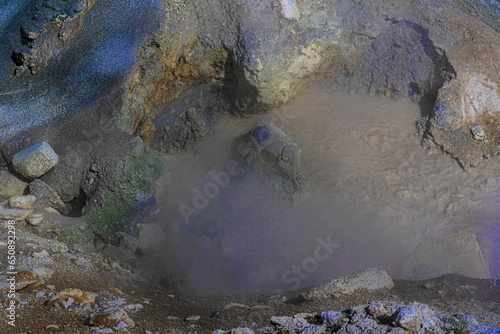 Top view shows mud surface surrounded with volcanic mineral rock and sediment in colors. Ground slopes to the watery muddy pool with bubbling muck in Yellowstone National Park.