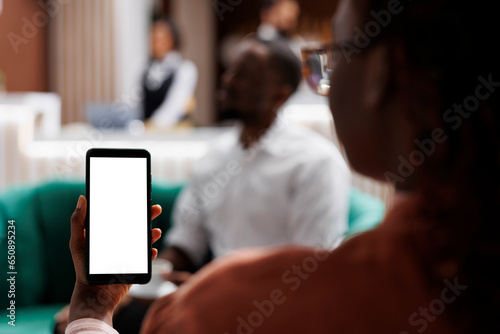 Tourist holding device with white screen, relaxing on sofa in lounge area at luxury hotel. Young adult guest using smartphone with blank display and isolated chromakey in lobby.