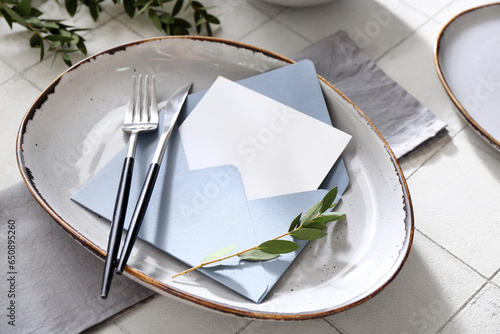 Stylish table setting, envelope with blank card and plant branches on white tile background