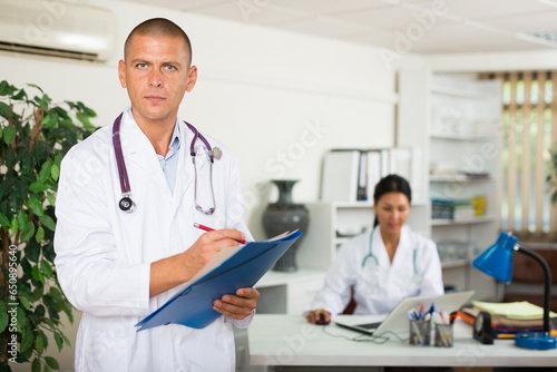 Positive male doctor standing in modern office with documents  writing medical history sheet