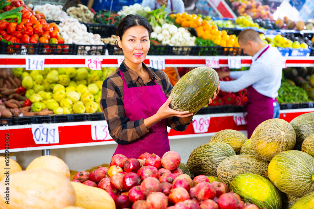 Asian woman in uniform standing in greengrocer and holding melon in hands. Her male colleague working in background.