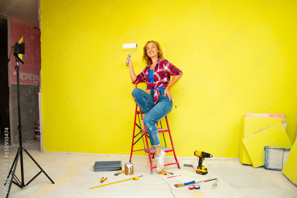 A beautiful young woman n a red plaid shirt, blue jeans and white sneakers  sits on a red stepladder with a roller in her hand against the background of a bright yellow wall