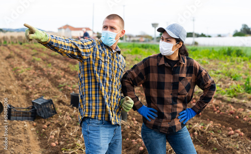 Young man and woman farm workers in masks having discussion at a field with potatoes, pointing with finger to something