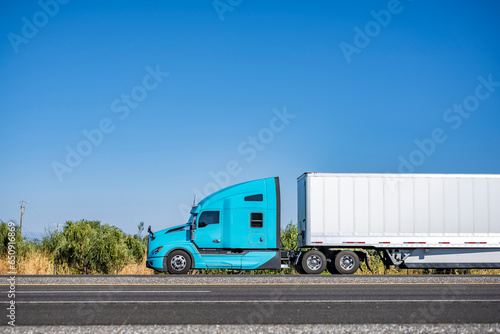 Turquoise big rig long hauler semi truck carrier transporting cargo in dry van semi trailer driving on the flat highway road in California