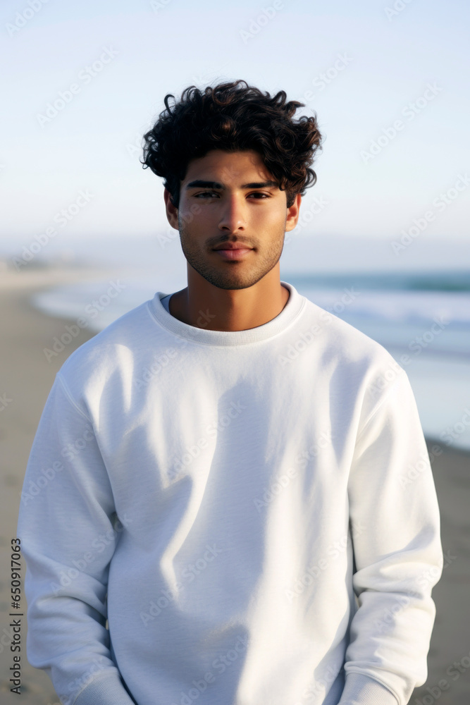 Young Hispanic man posing in a white crewneck, looking at the camera, against a softly focused beach shoreline. Young Hispanic man posing in a white crewneck, looking at the camera, against a softly focused beach shoreline.