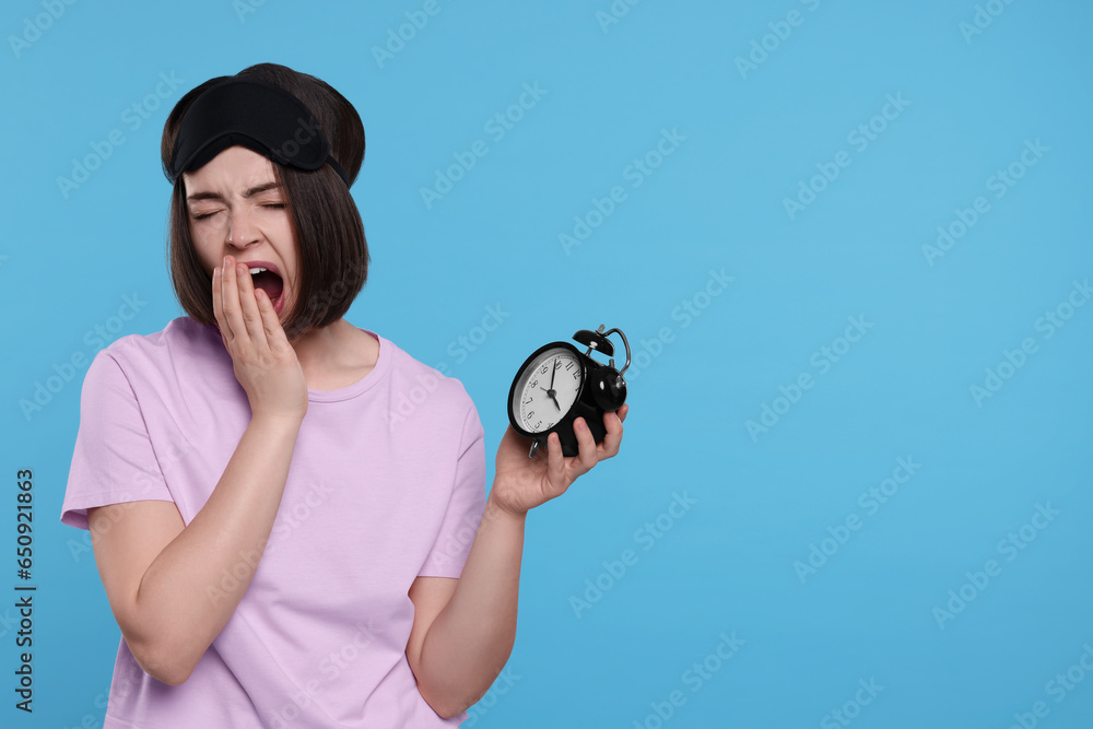 Fototapeta premium Sleepy young woman with sleep mask and alarm clock yawning on light blue background, space for text. Insomnia problem