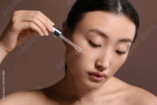 Beautiful young woman applying cosmetic serum onto her face on brown background, closeup