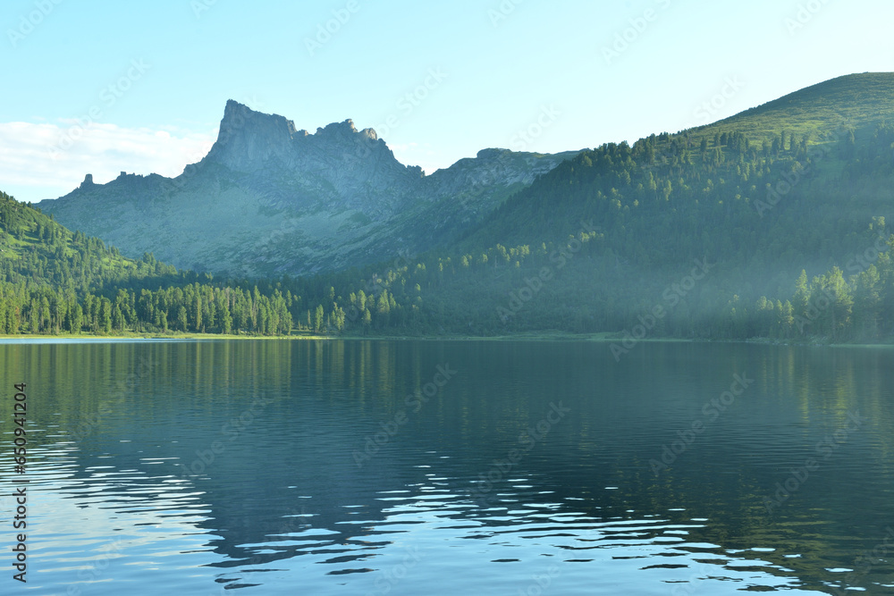 A light haze over the unsteady surface of a large lake in a dense coniferous forest at the foot of high peaked mountains.