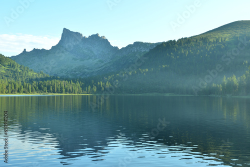 A light haze over the unsteady surface of a large lake in a dense coniferous forest at the foot of high peaked mountains.